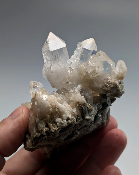 A clear quartz crystal cluster with pointed, transparent spikes emerging from a rough, grayish rock base.