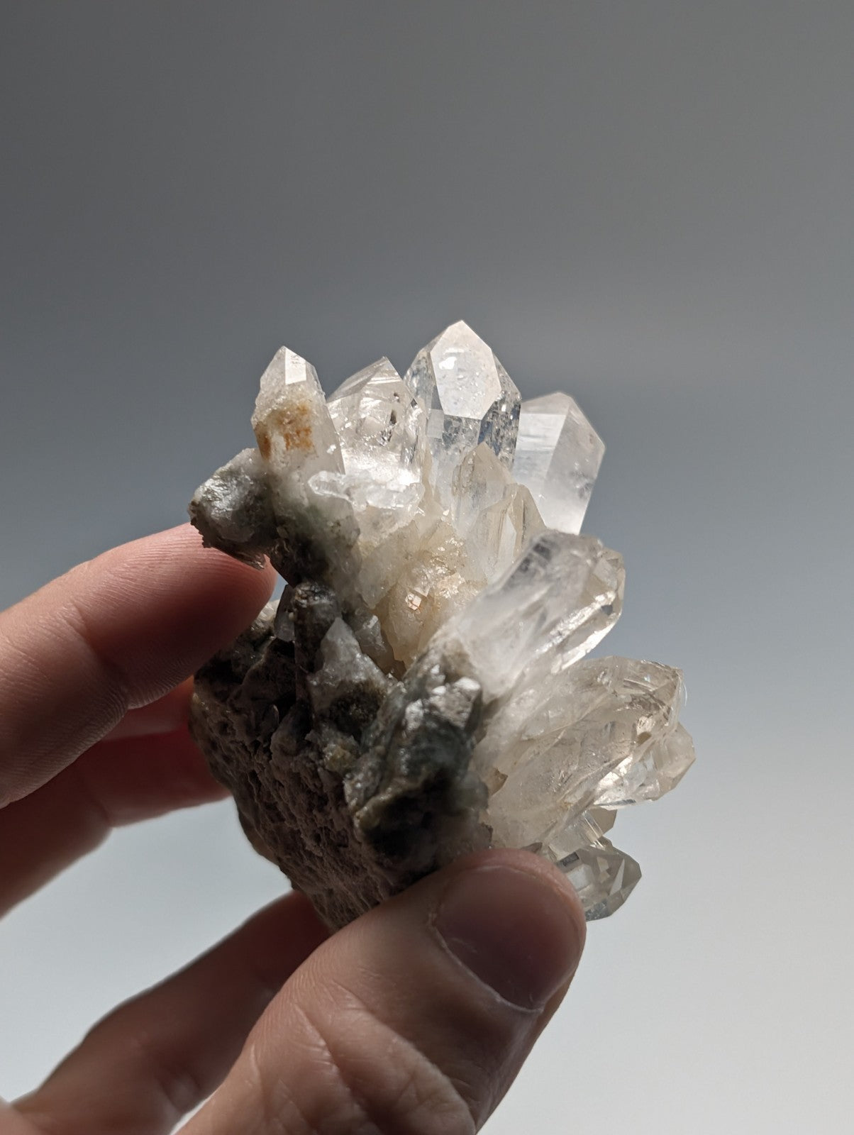 A cluster of clear, faceted quartz crystals growing from a dark, porous rock base.