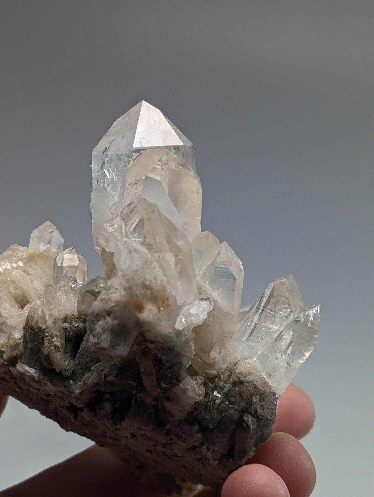 A clear quartz crystal cluster with a prominent, sharp-pointed central spike and smaller surrounding crystals growing from a dark gray rock base.