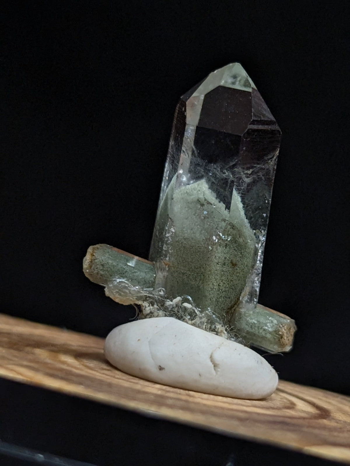 A translucent green quartz crystal cluster with a pointed top and rough, mossy-green base rests on a smooth white pebble.