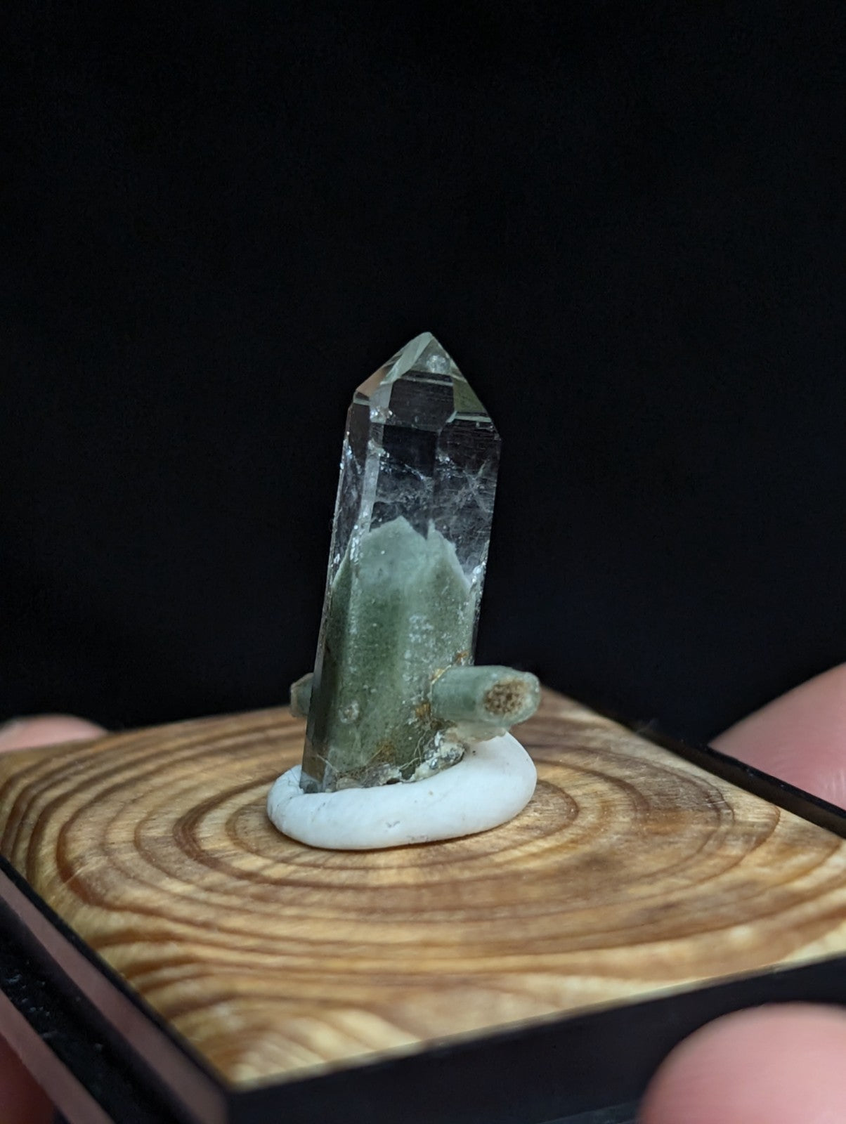 A clear quartz crystal with a greenish base and natural inclusions, displayed on a white resin stand atop a wooden plinth.