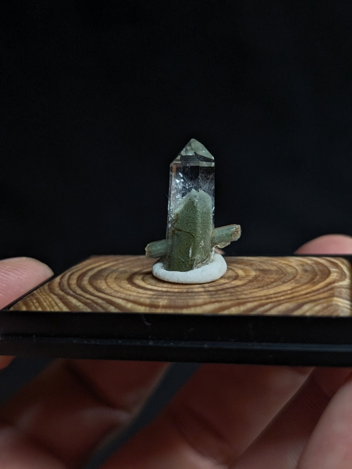 A clear quartz crystal with greenish base and branching formations, mounted on a white circular pedestal atop a wooden display board.