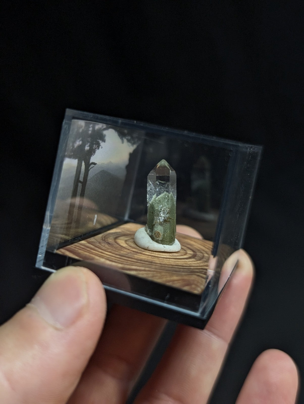 A translucent green crystal with a pointed top and natural inclusions, displayed inside a clear acrylic box on a wooden base.