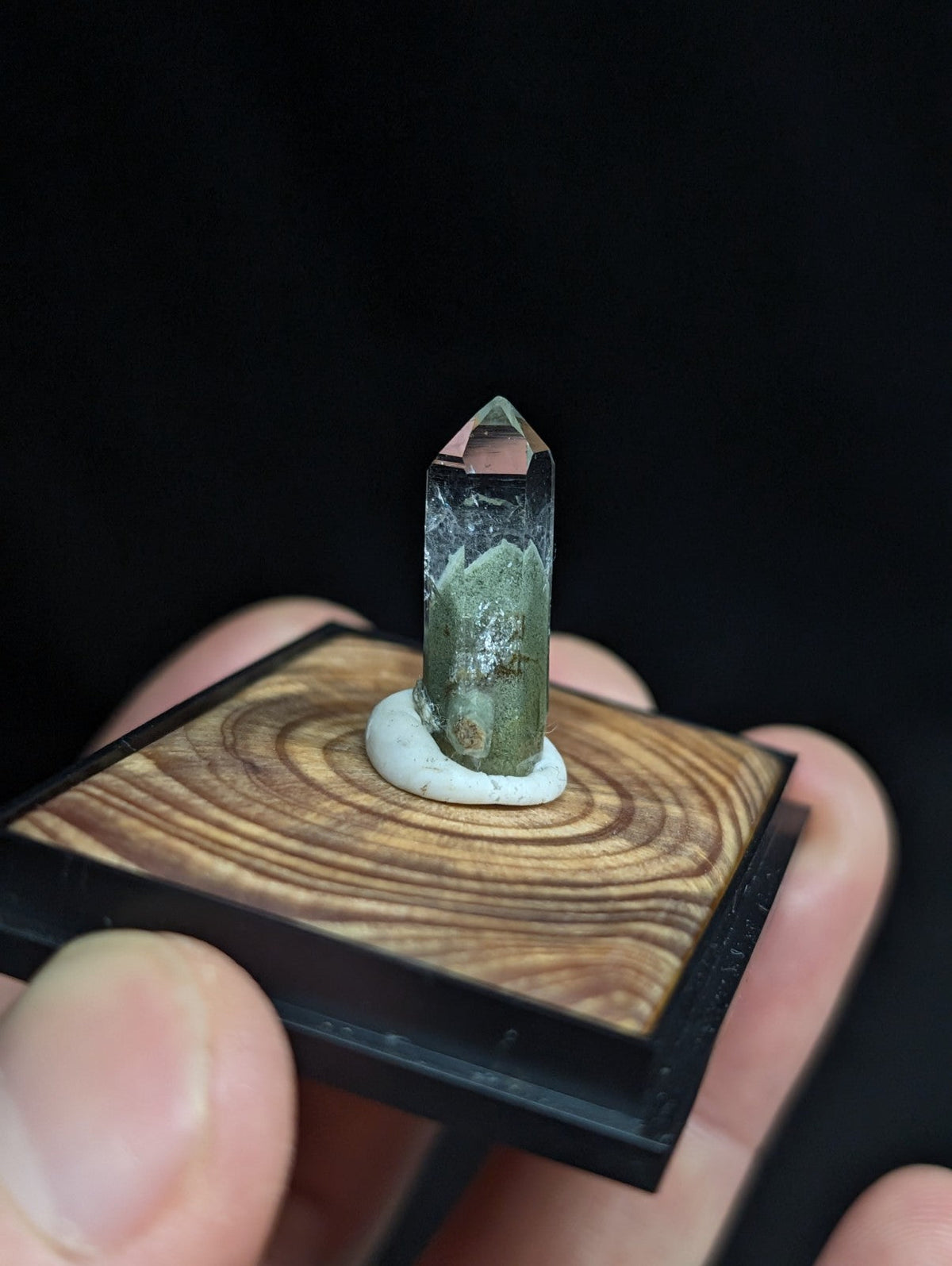 A translucent green crystal with a pointed top, mounted on a circular white base and set within a square wooden display stand.