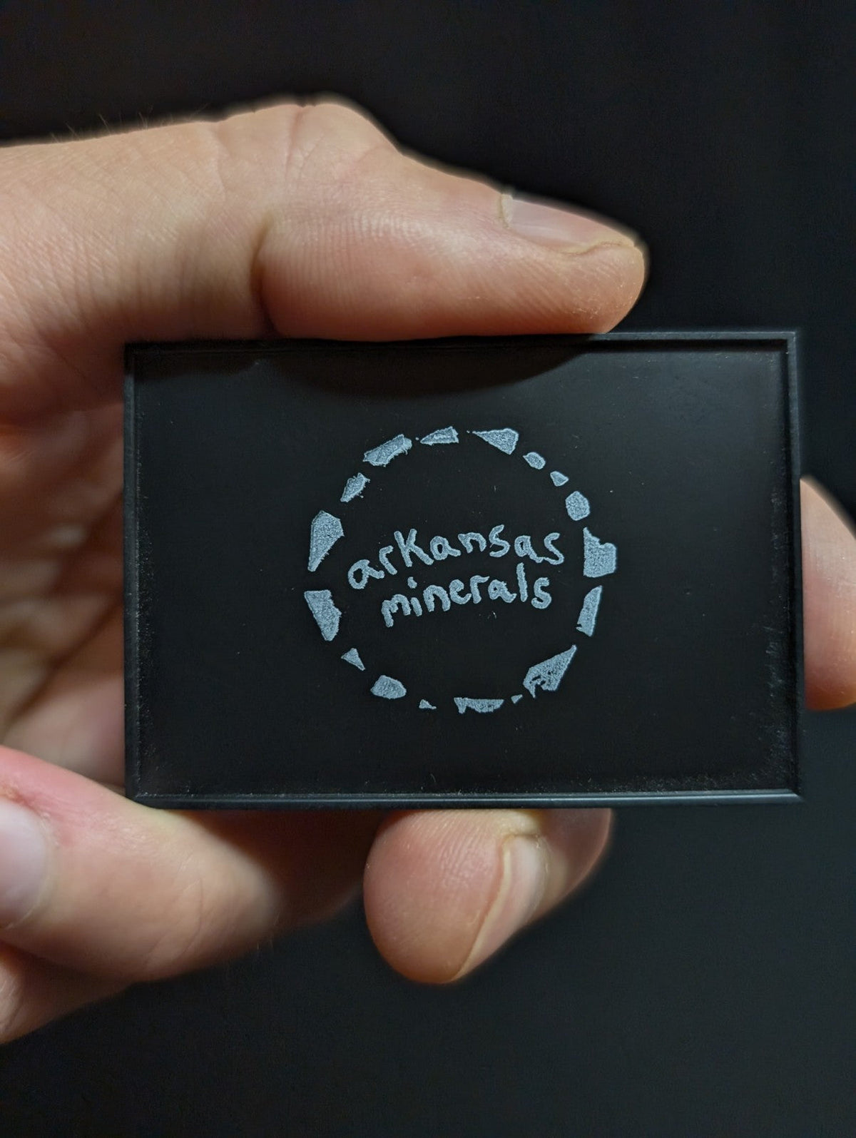 Black rectangular box with silver ’arkansas minerals’ logo encircled by a ring of stylized mineral shapes.