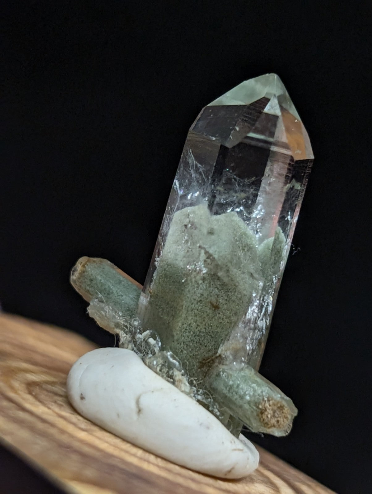 A clear quartz crystal with greenish inclusions and a pointed tip, mounted on a white stone base.