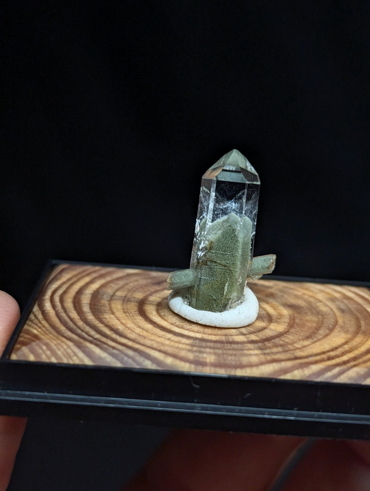 A translucent green crystal with a pointed top and natural inclusions, mounted on a white circular base atop a wooden display board.