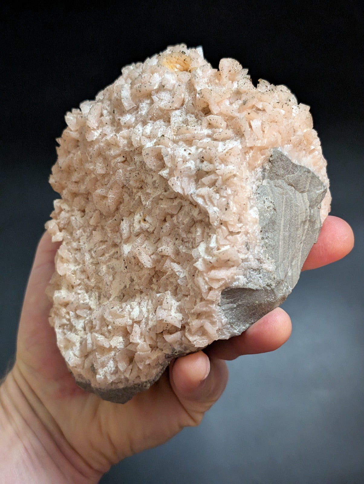 A hand holds a pale pink, crystalline mineral specimen with a rough, textured surface and some darker gray rock matrix.