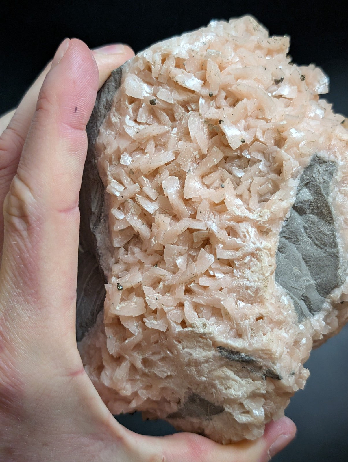 A hand holds a rough, pale pink mineral specimen with translucent, crystalline formations growing from a darker gray rock base.