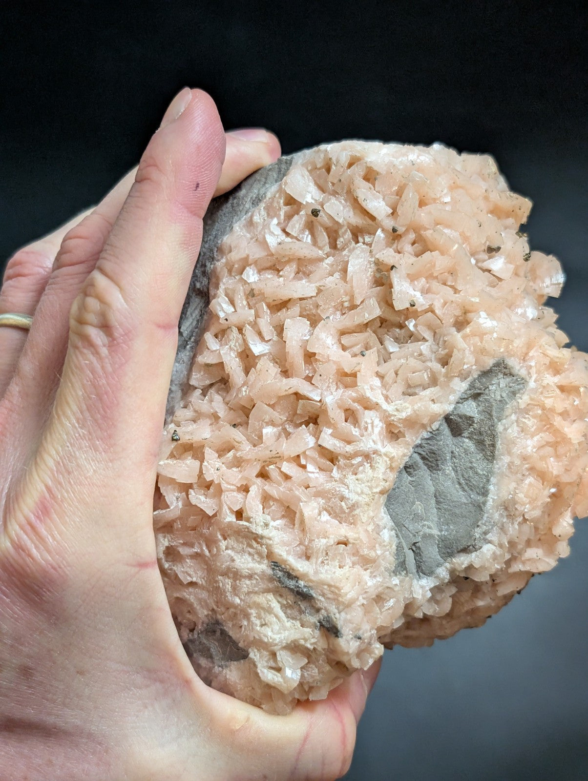 A hand holds a rough, pinkish mineral specimen with crystalline formations and dark gray matrix.