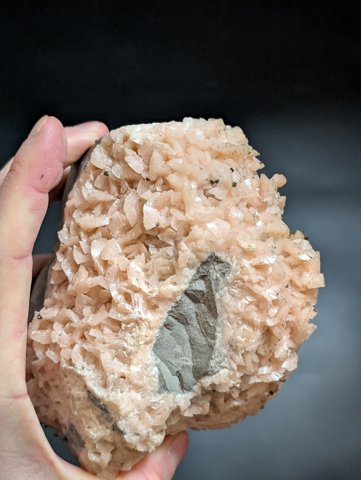 A hand holds a rough, pinkish-orange mineral specimen with crystalline formations and a dark gray interior cavity.