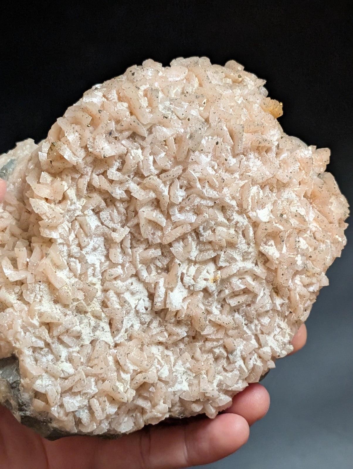 A hand holds a pale pink, crystalline mineral specimen with intricate, interlocking facets and subtle speckles of darker material.