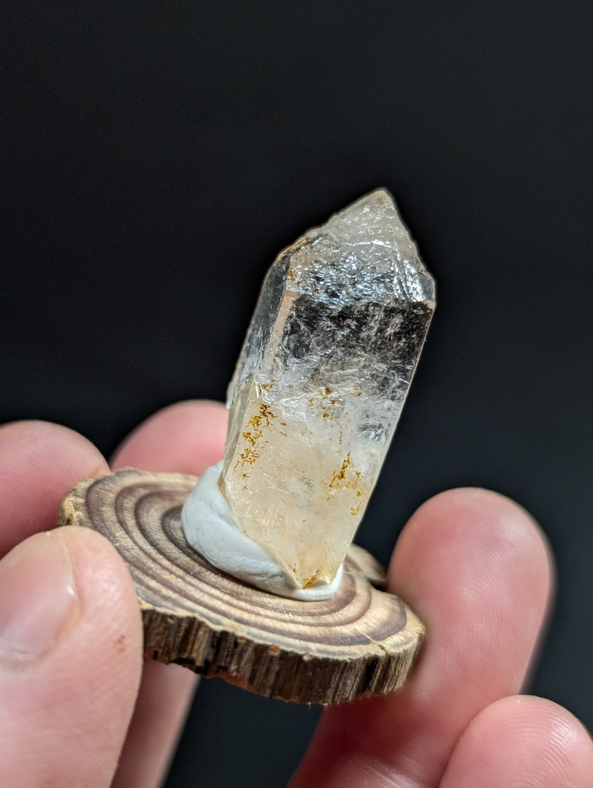 A translucent, clear quartz crystal with golden inclusions and a pointed tip, mounted on a rustic wooden slice.