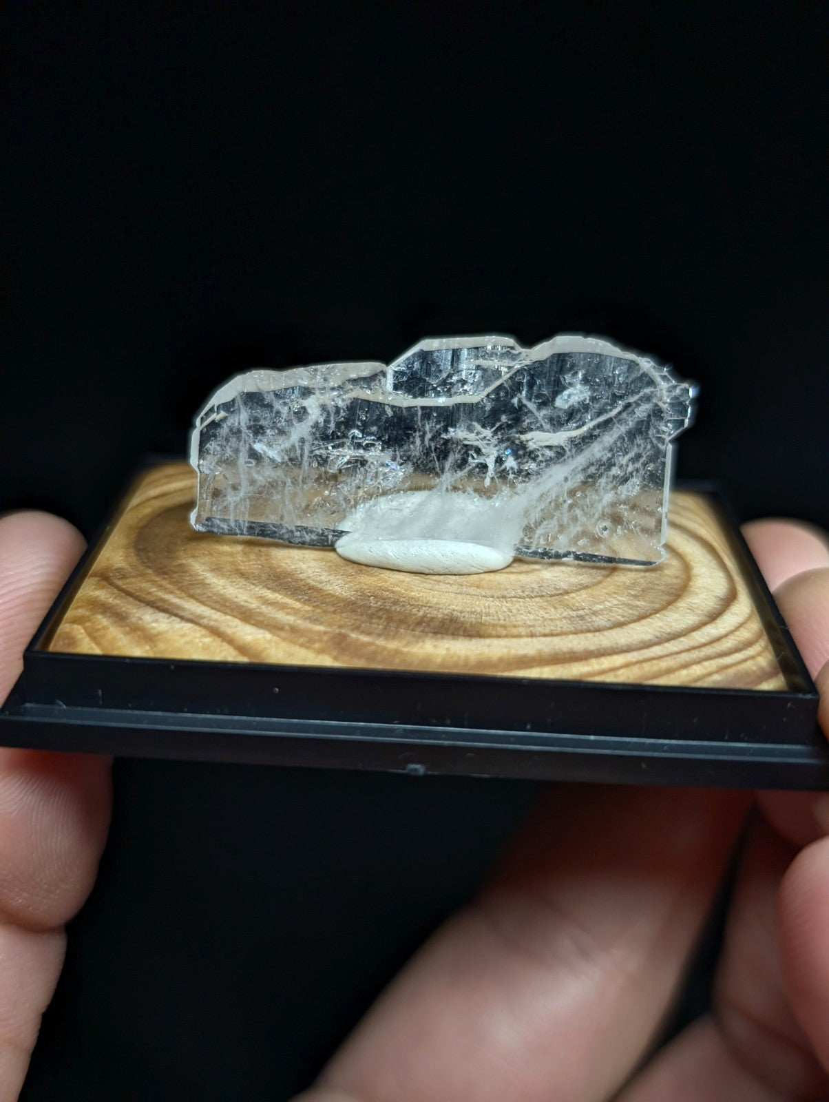 A translucent, clear quartz crystal with a rough, jagged surface and an embedded white mineral inclusion rests on a wooden display base.