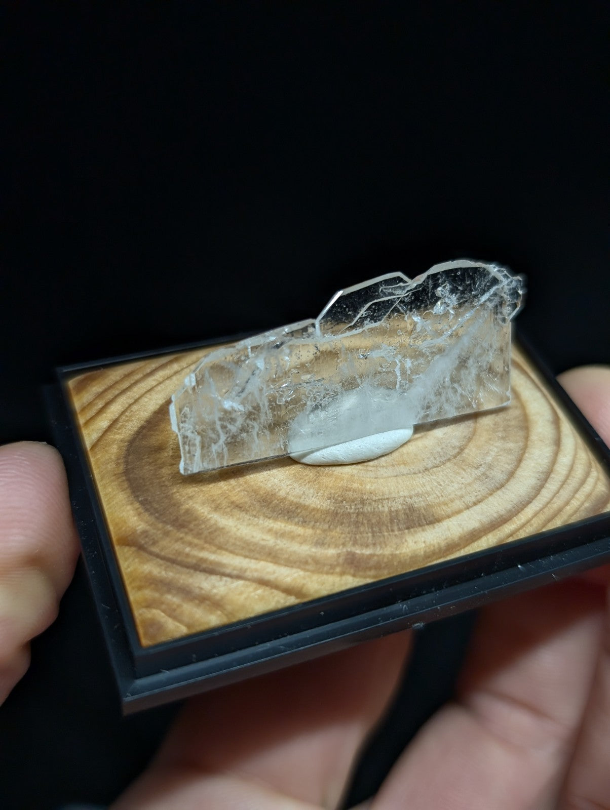 A translucent, clear quartz crystal with natural fractures and a smooth base rests in a black-framed wooden display case.