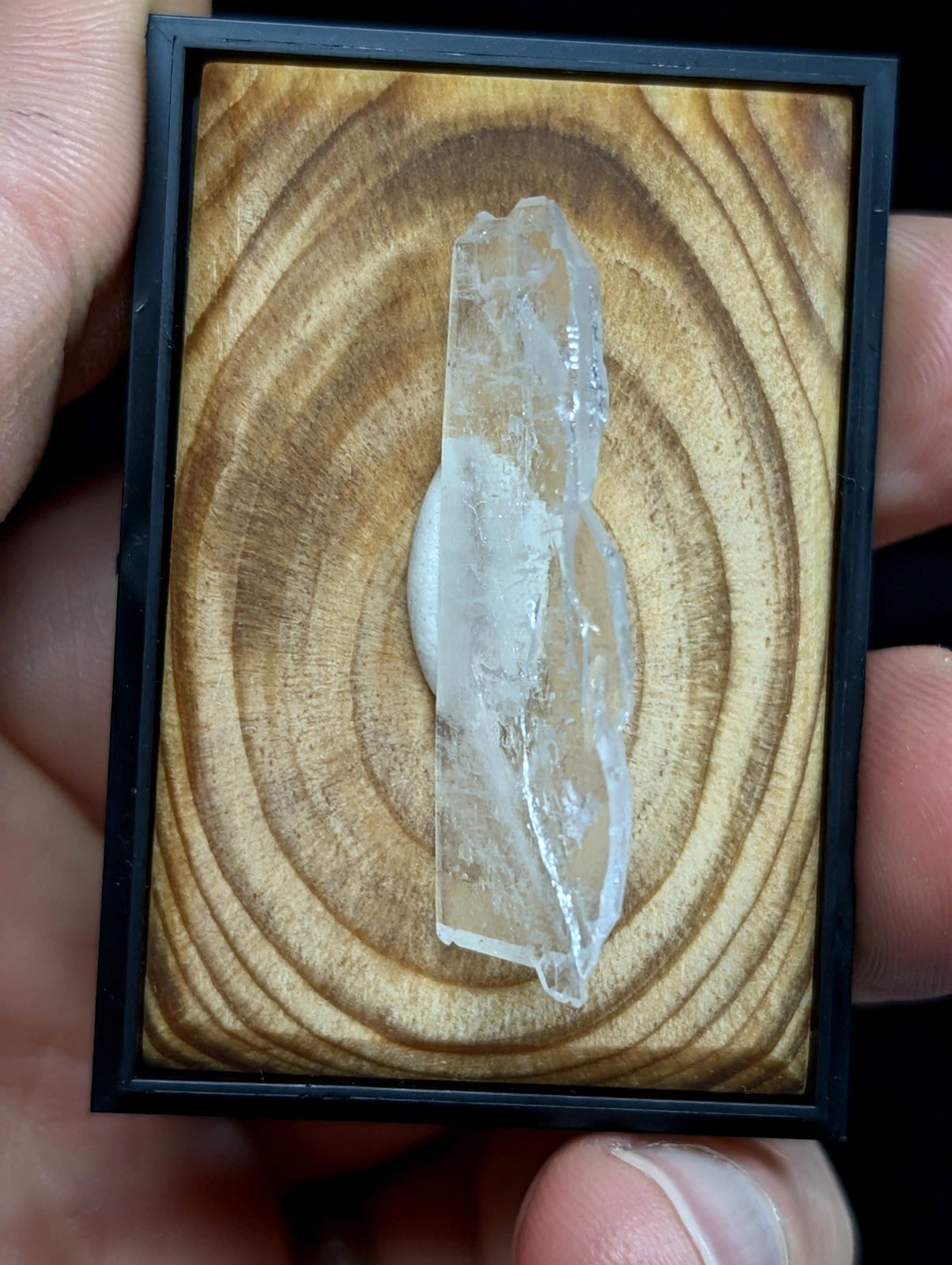 A clear, translucent quartz crystal with natural facets and a slightly cloudy base is displayed in a black-framed wooden panel.