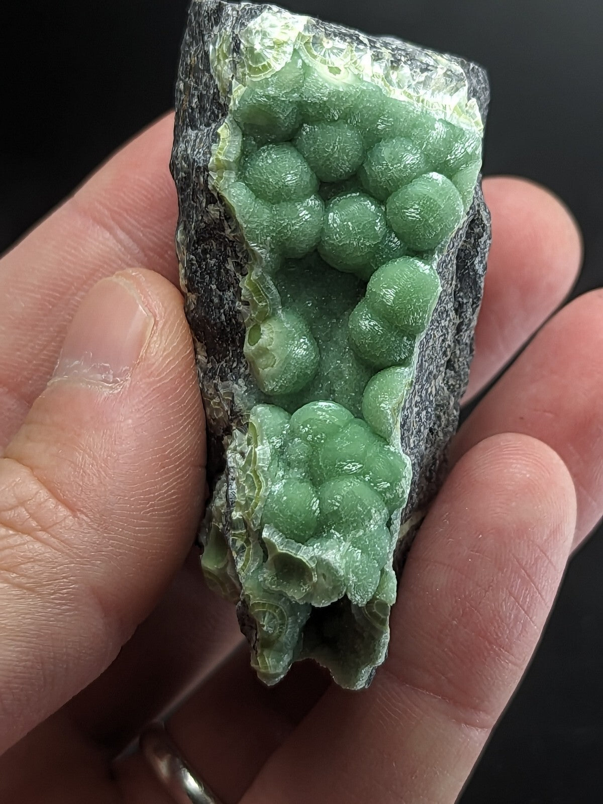 A hand holds a geode with vibrant green, spherical crystals growing from a dark gray rock matrix.
