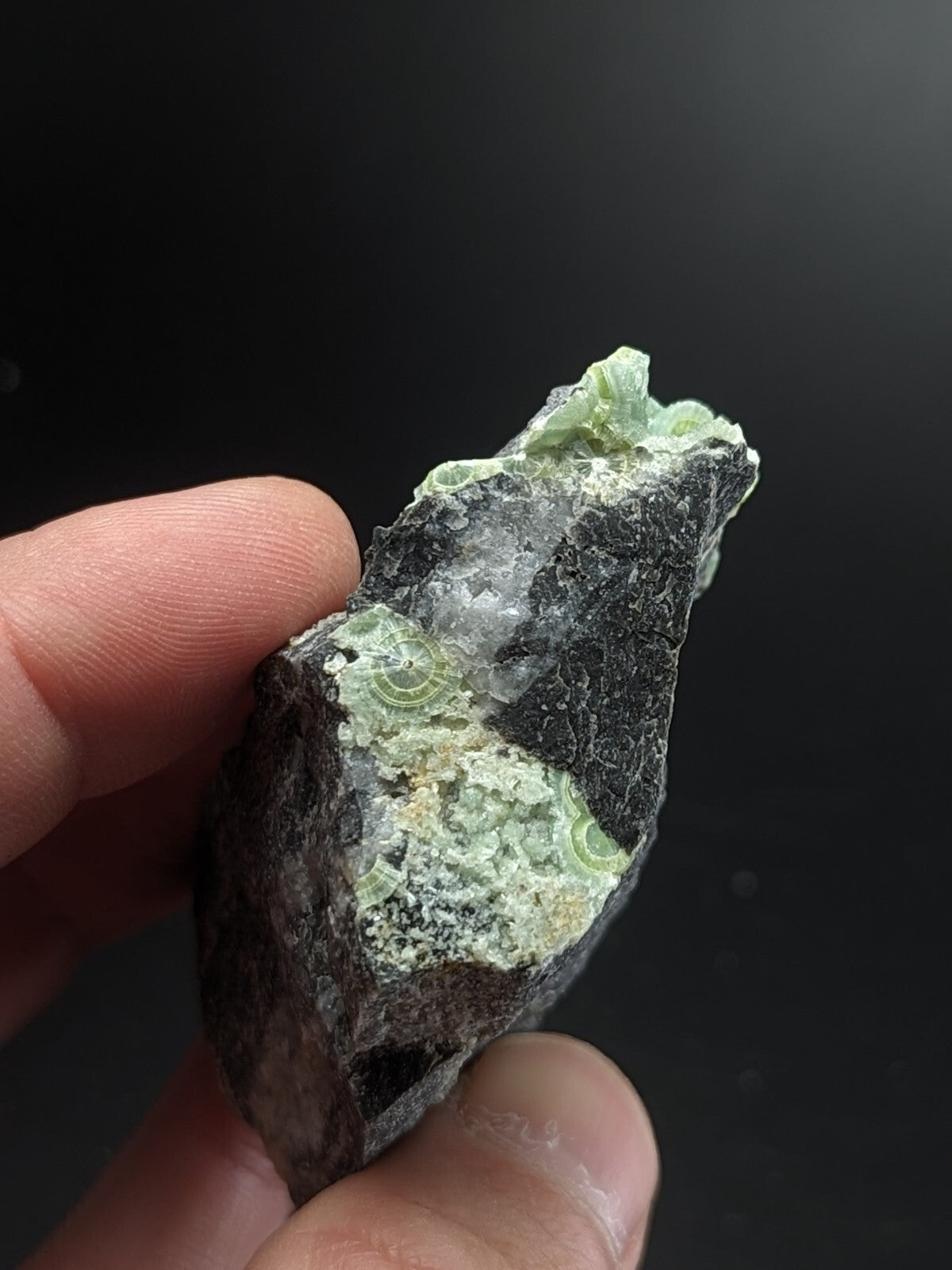 A hand holds a rough, irregularly shaped mineral specimen featuring dark gray rock with patches of translucent green and white crystalline formations.