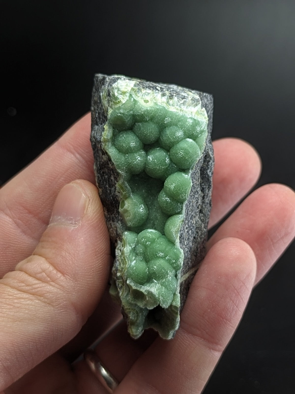 A hand holds a dark gray rock with vibrant green, spherical crystals growing from its surface.