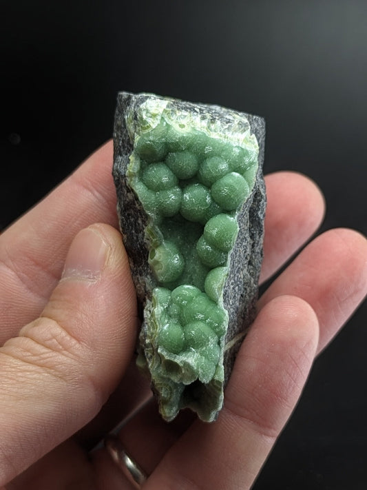 A hand holds a dark gray rock with vibrant green, spherical crystals growing from its surface.