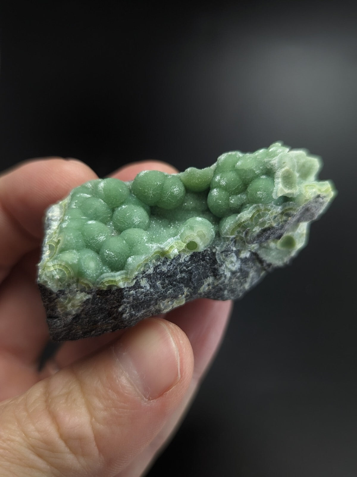A hand holds a piece of green, spherical mineral formation with a rough, dark gray base.