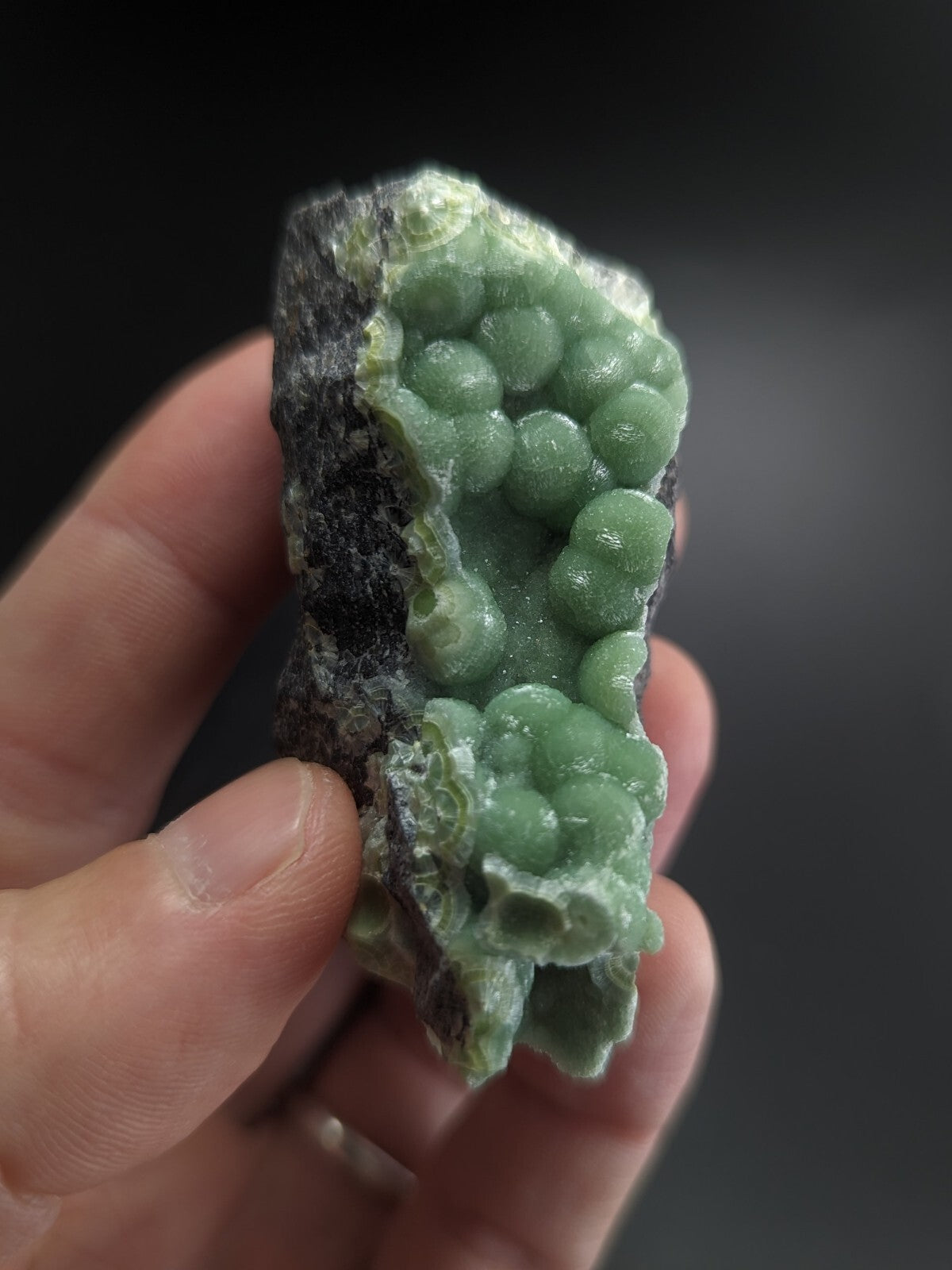 A hand holds a geode with translucent green spherical crystals growing from a dark, rough-textured rock base.