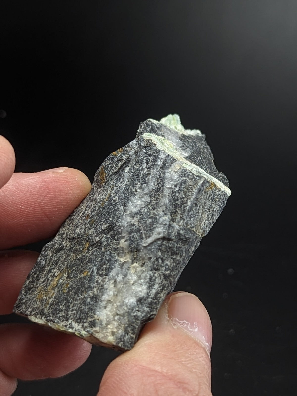 A rugged, dark gray rock specimen with streaks of white and faint green mineral veins.