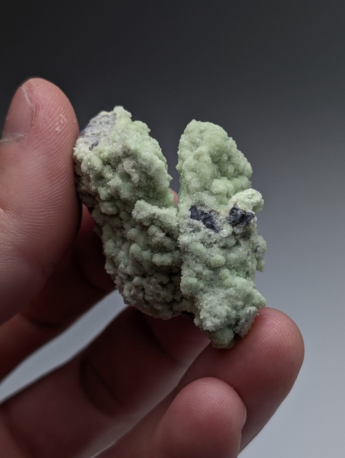A pale green fluorwavellite mineral with dark inclusions intermixed with SiO2 held between fingers from Mauldin Mountain Arkansas