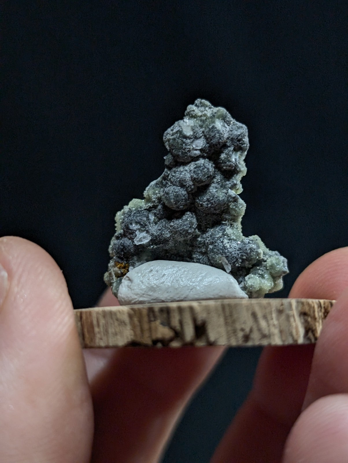 A textured, grayish-green mineral specimen with clustered crystalline formations rests on a natural wood display base.