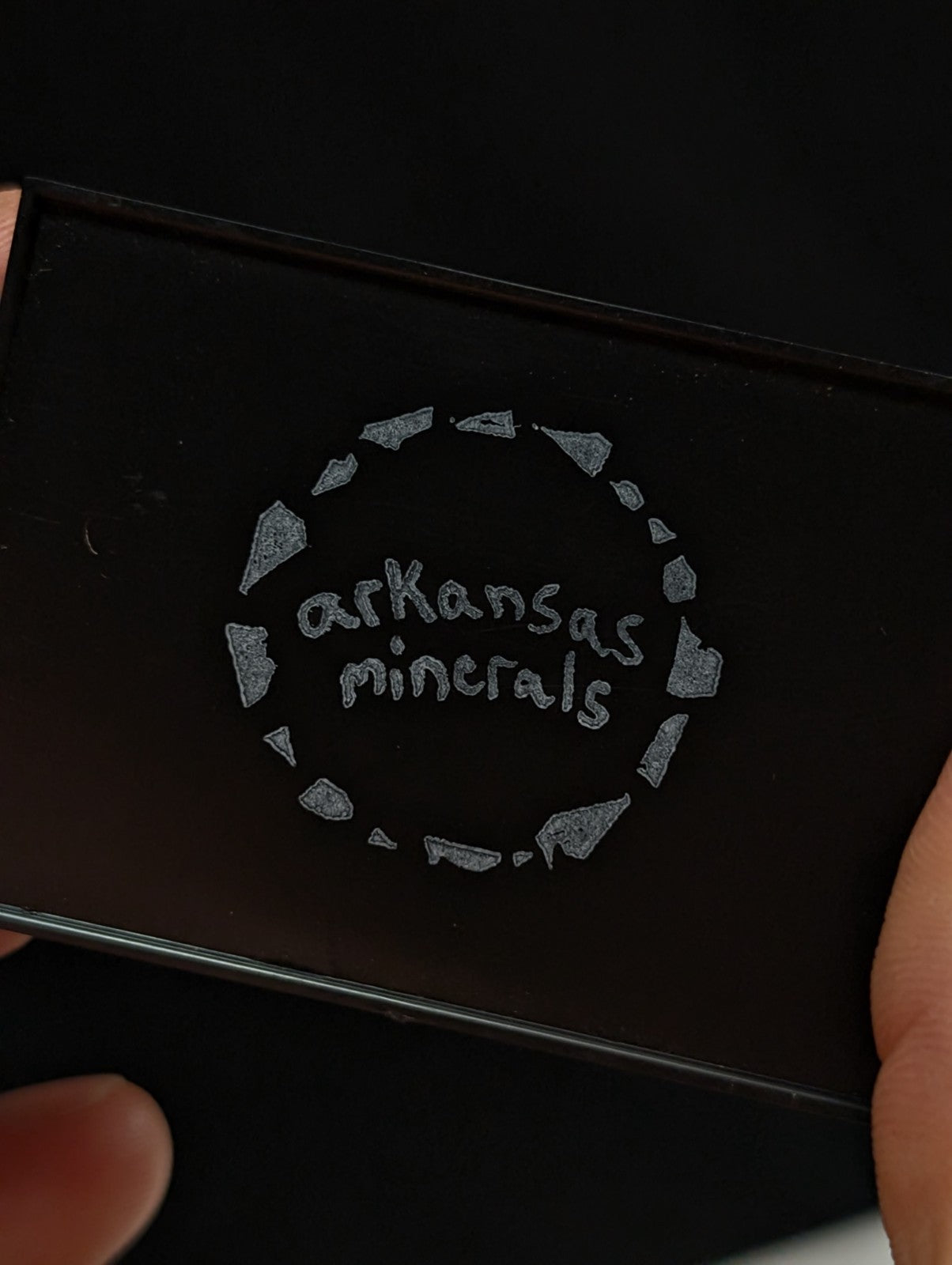 Black rectangular compact with a silver embossed ’arkansas minerals’ logo encircled by fragmented stone shapes.