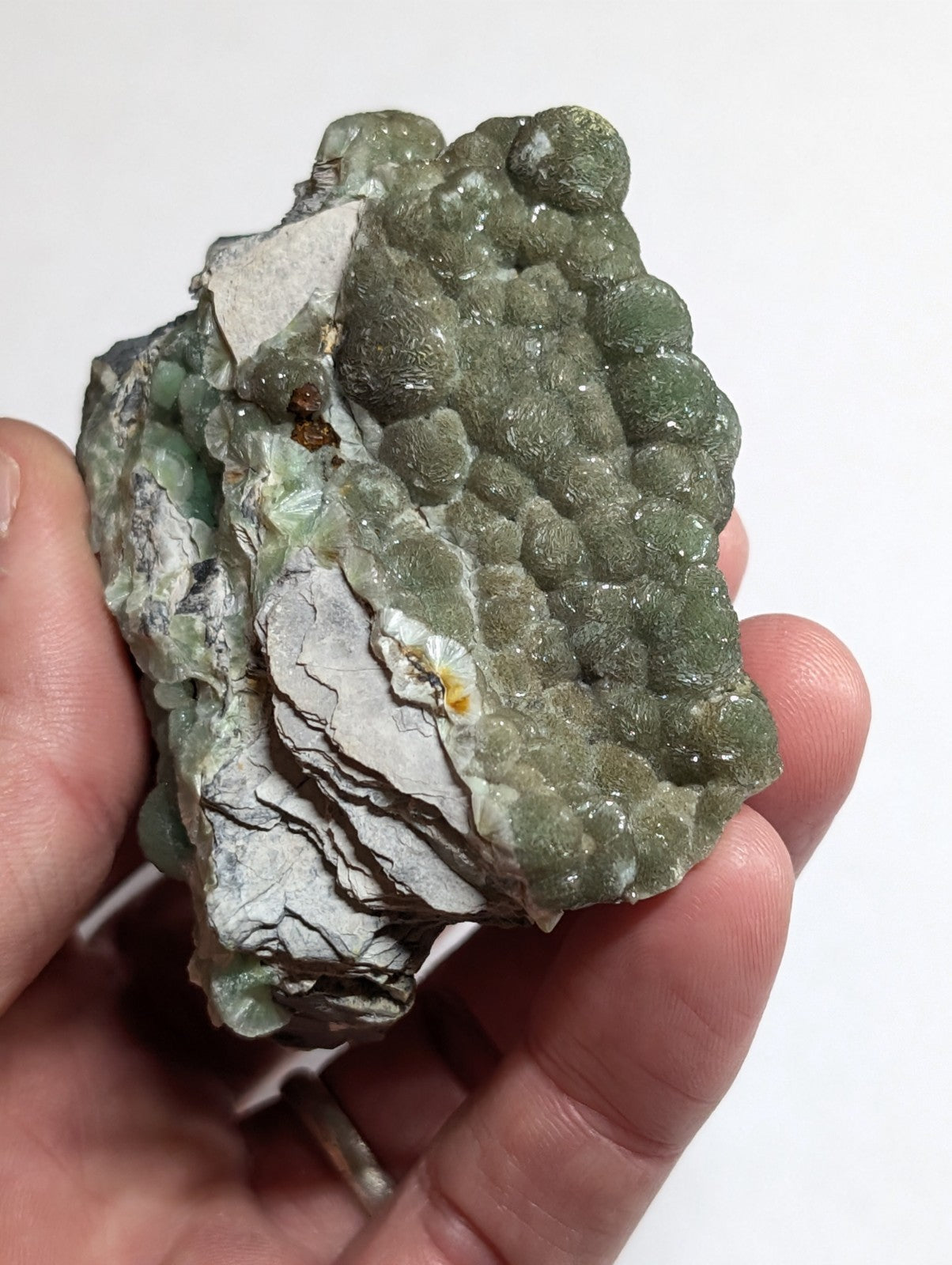 A hand holds a rough, irregularly shaped mineral specimen featuring clusters of translucent green crystals growing from a grayish matrix.