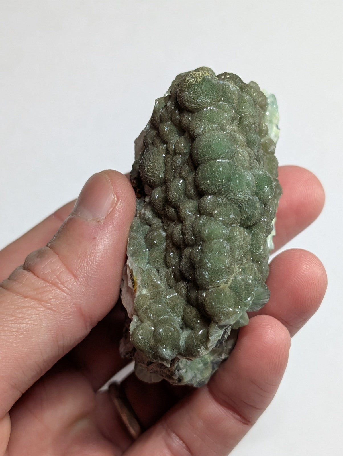 A hand holds a green, bumpy mineral specimen with rounded growths and translucent patches.