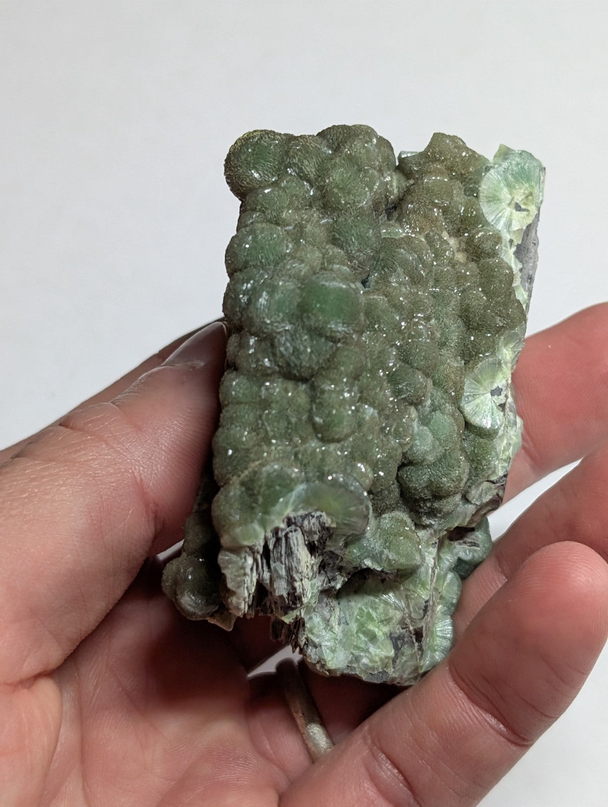 A hand holds a green, bumpy mineral specimen with rounded crystalline formations and some translucent patches.