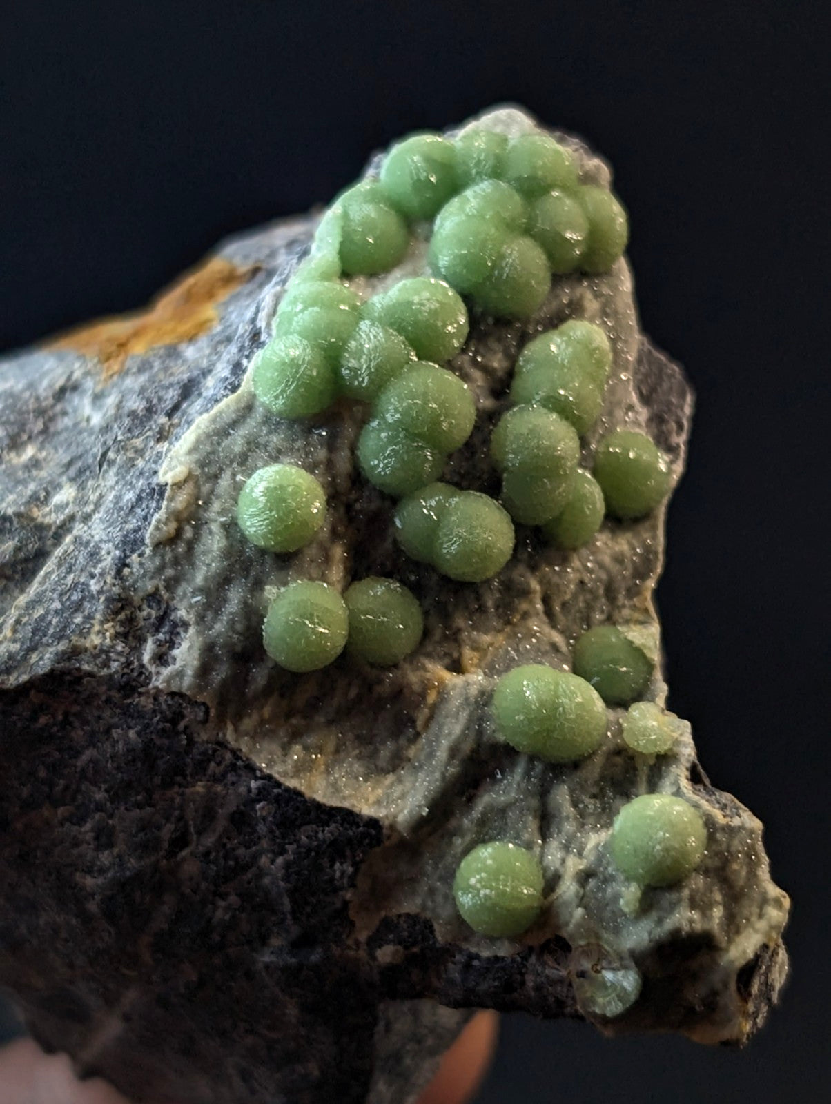 A cluster of translucent, light green spherical crystals grows naturally on a textured gray rock surface.