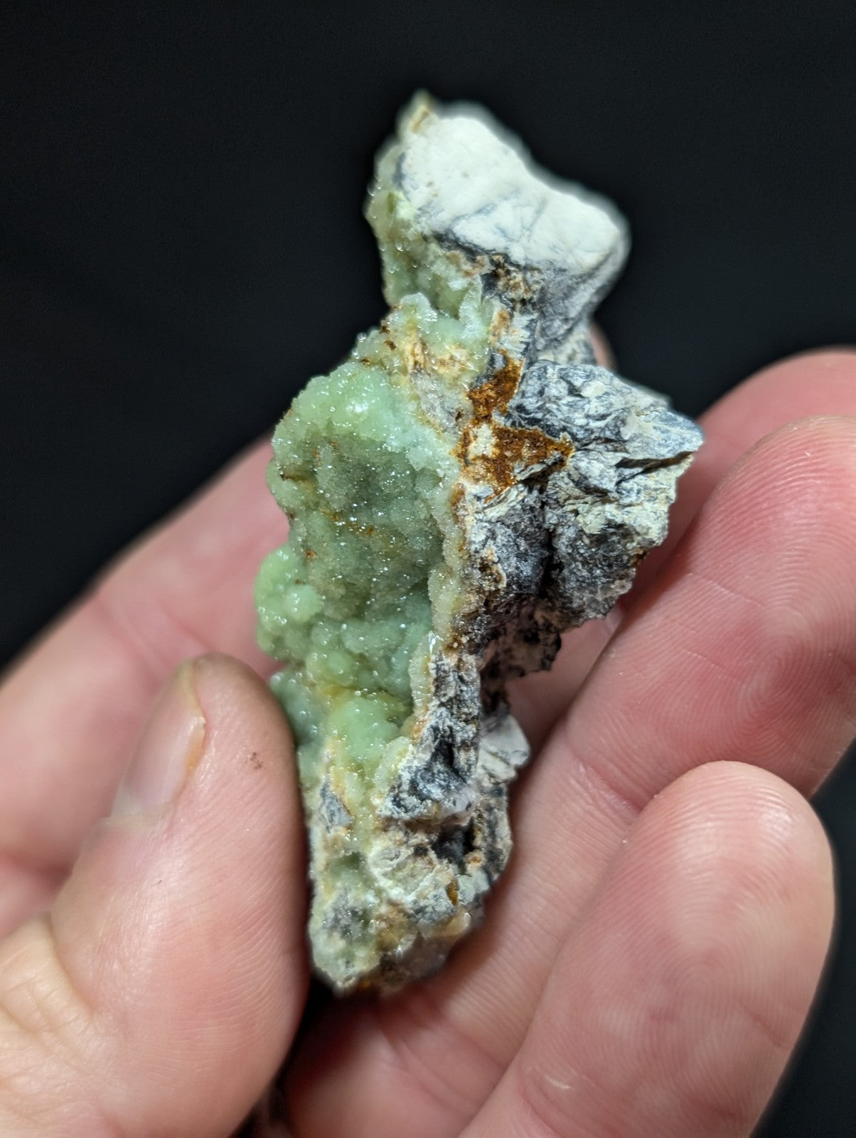 A hand holds a rough, irregularly shaped mineral specimen featuring vibrant green crystalline formations alongside grayish rock matrix and patches of rust-colored oxidation.