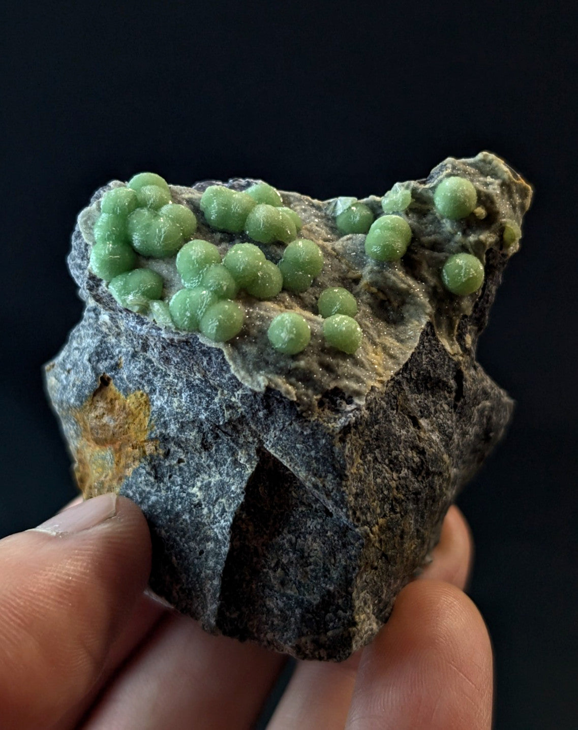 A hand holds a dark gray rock specimen featuring clusters of vibrant green, spherical crystals.