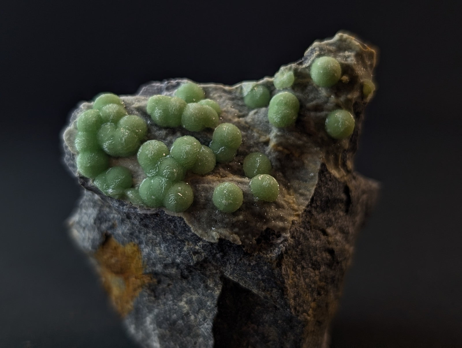A cluster of translucent, light green spherical crystals grows from a rugged gray rock matrix.