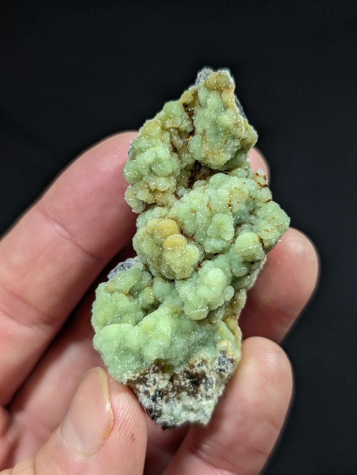 A hand holds a cluster of pale green, crystalline mineral formations with hints of yellow and dark matrix.