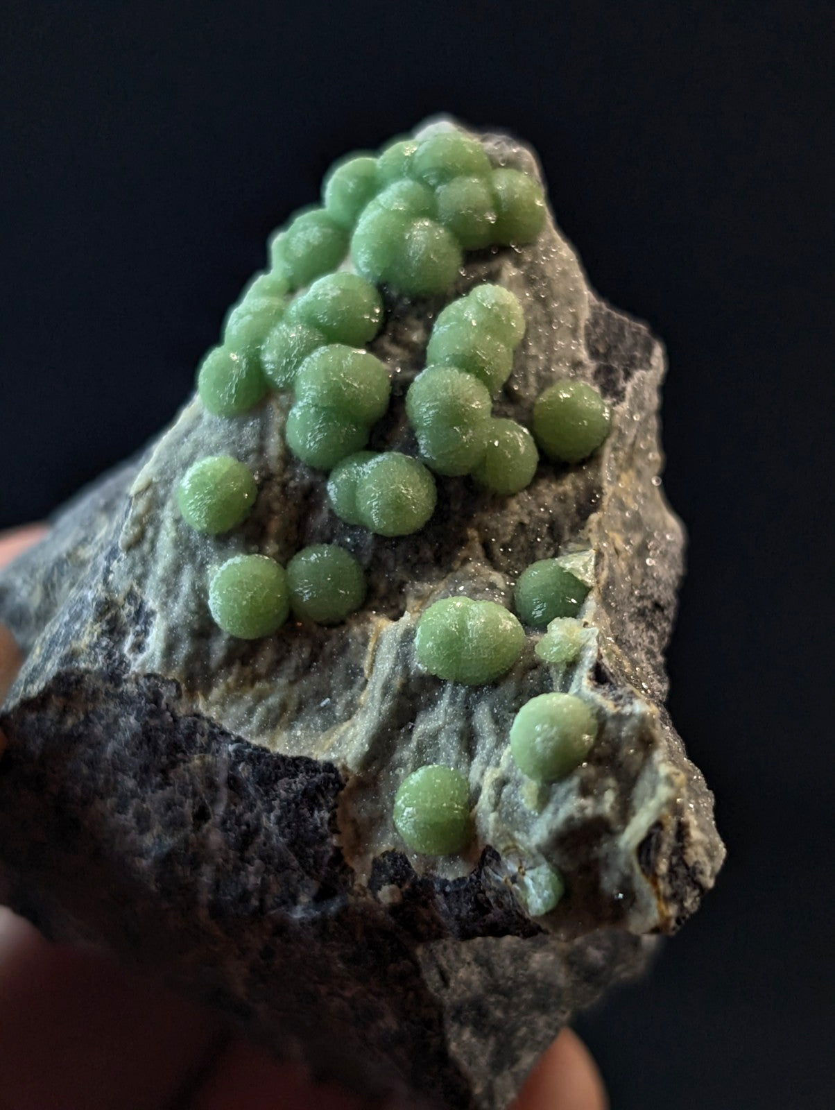 A cluster of translucent, light green spherical crystals grows densely on a rough-textured gray rock.