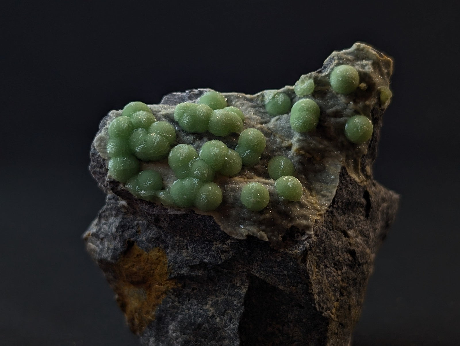 A cluster of translucent green spherical crystals growing on a dark, textured rock surface.