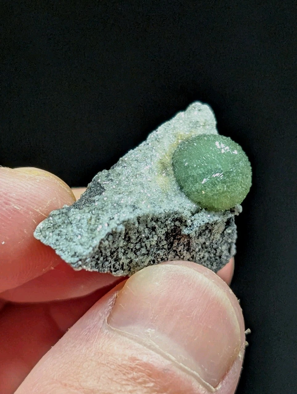 A green, translucent spherical mineral nestled atop a gray, speckled rock fragment.