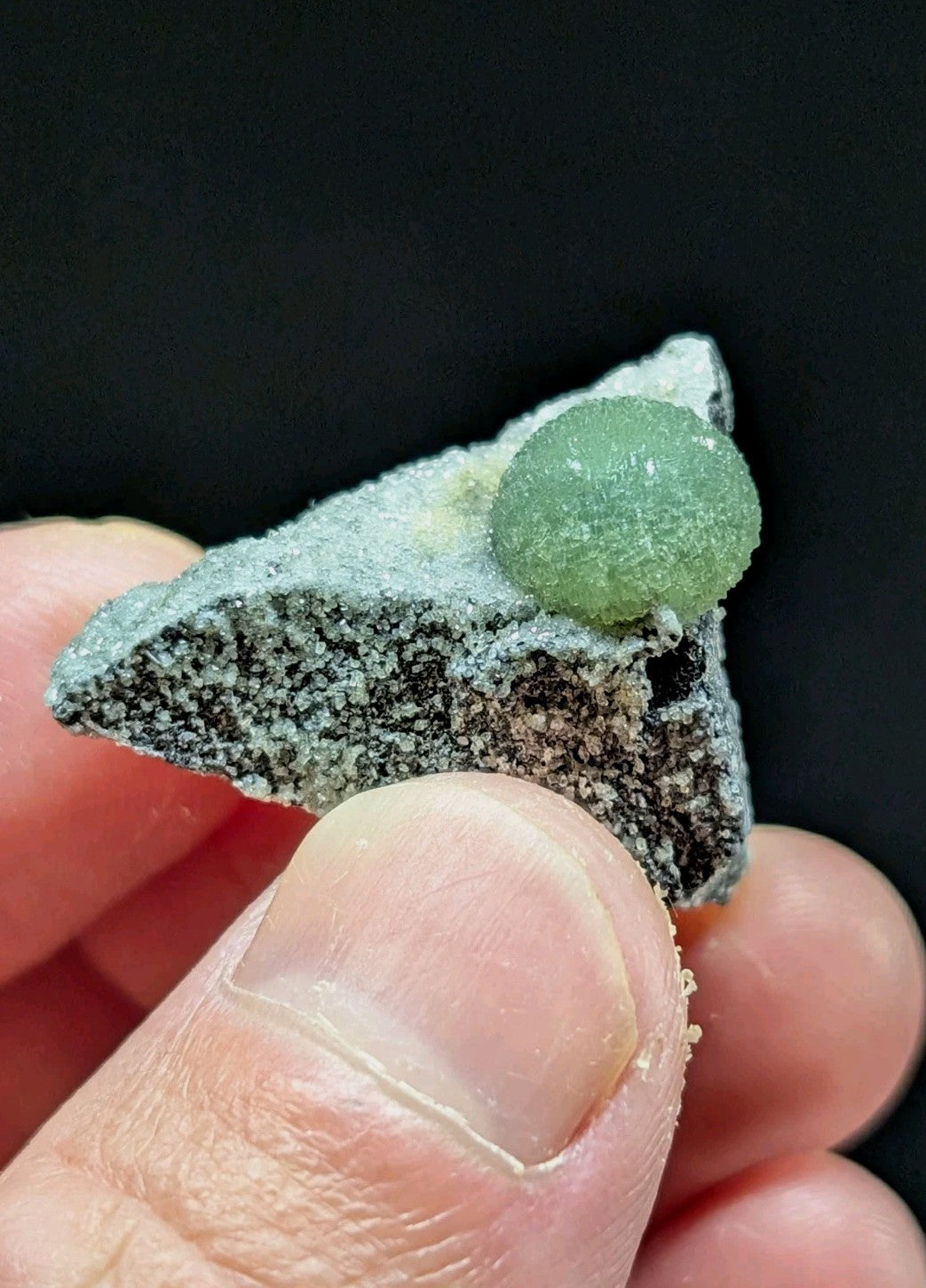 A translucent green spherical mineral specimen nestled atop a textured, speckled gray rock.