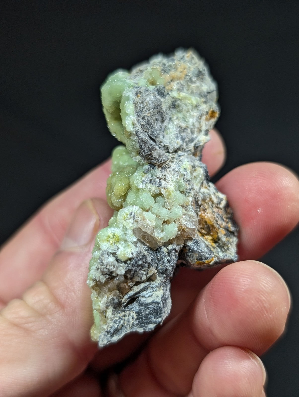A hand holds a rough, irregularly shaped mineral specimen featuring clusters of translucent green crystals embedded in a grayish matrix with patches of yellow and black.