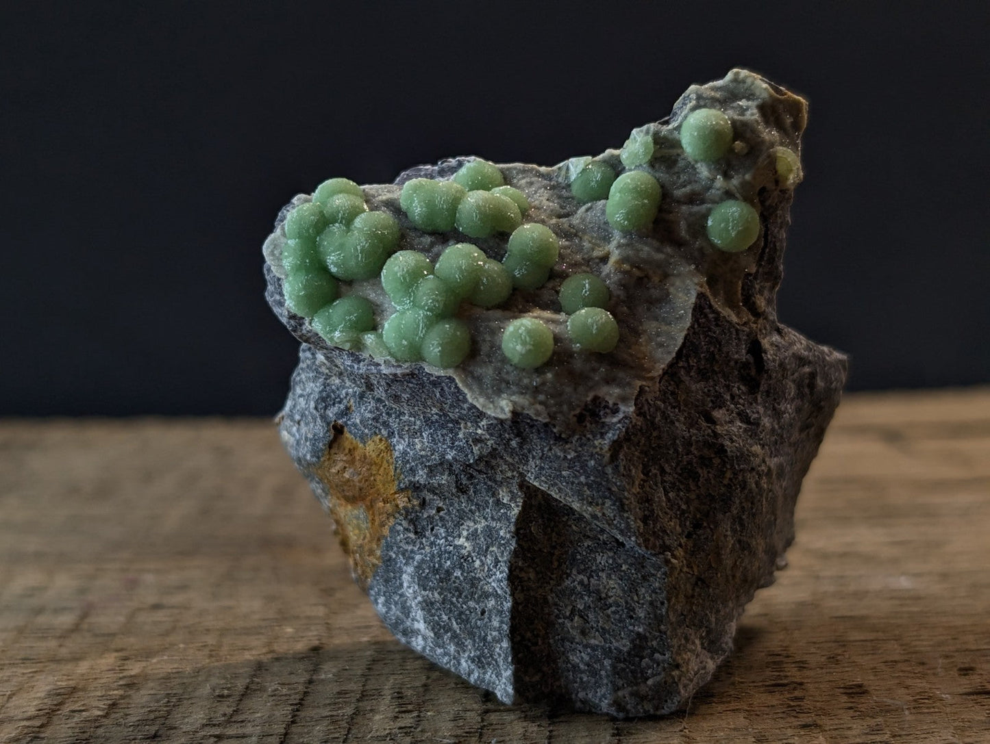 A cluster of vibrant green, spherical mineral formations grows atop a rugged gray rock with subtle golden flecks.