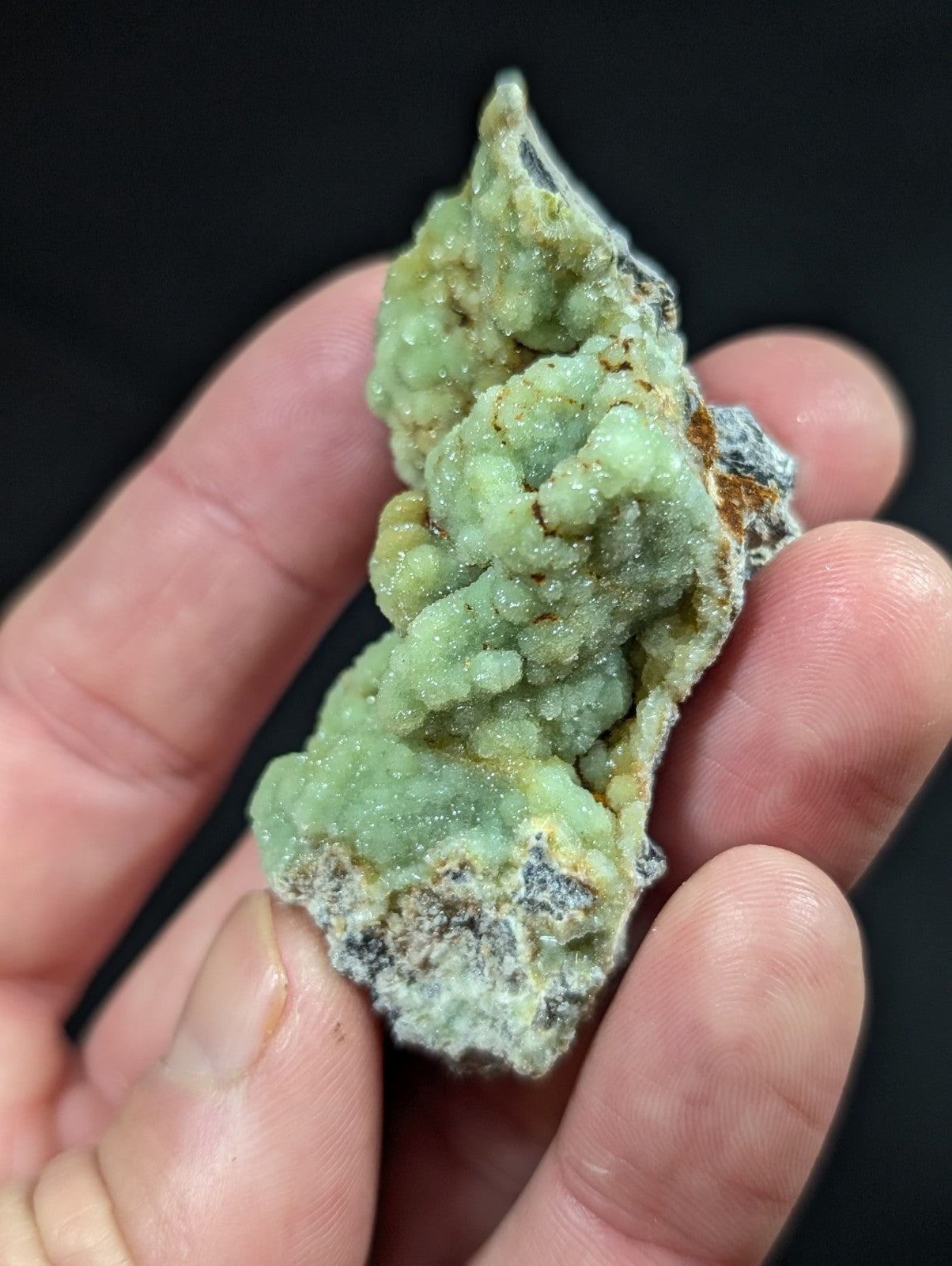 A hand holds a rough, light green mineral specimen with crystalline formations and patches of darker matrix.