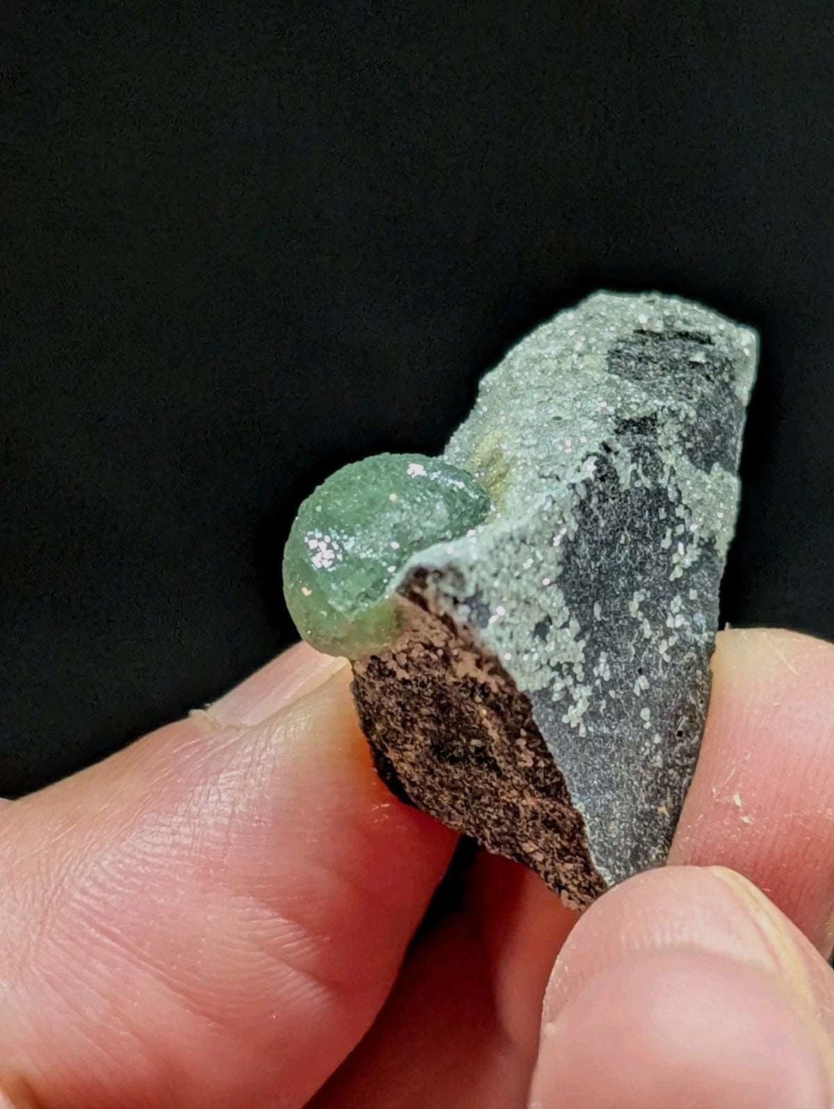 A translucent green gemstone nestled within a rough, speckled gray rock matrix.