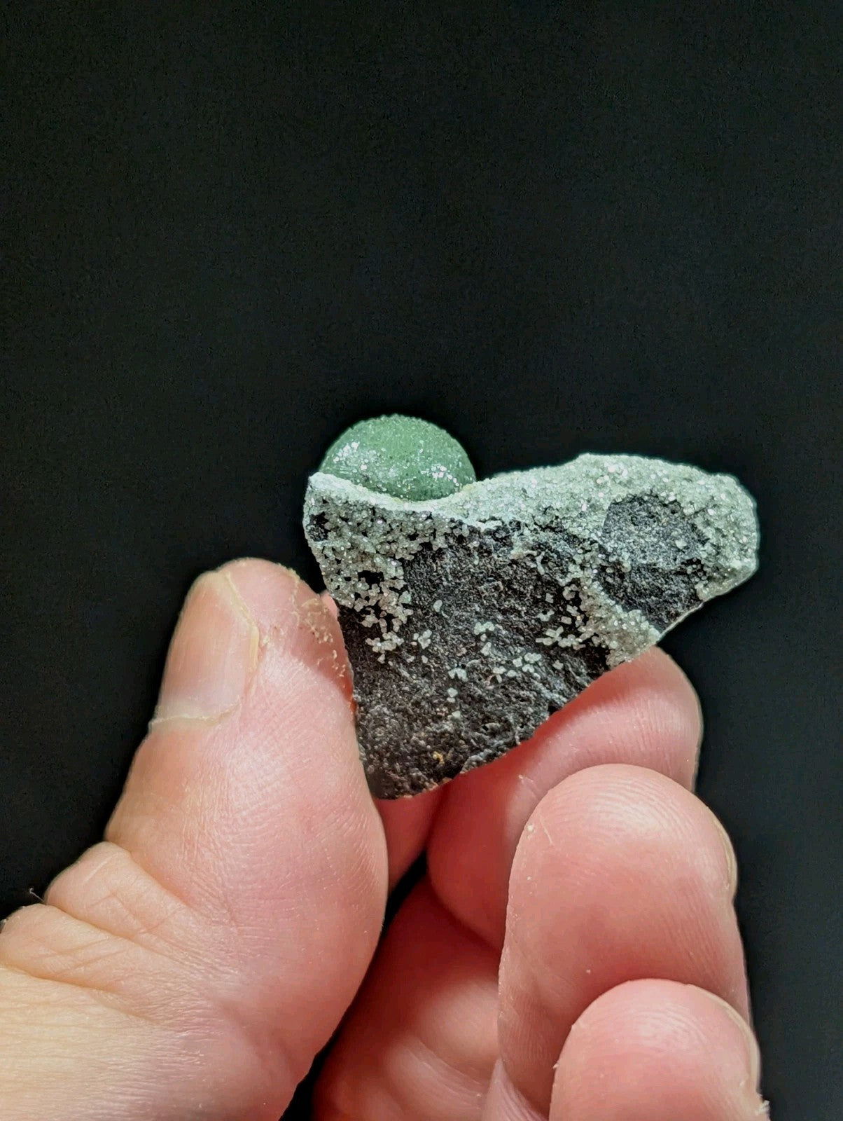 A hand holds a rough, grayish-green rock fragment with a smooth, spherical green gem nestled in its crevice.