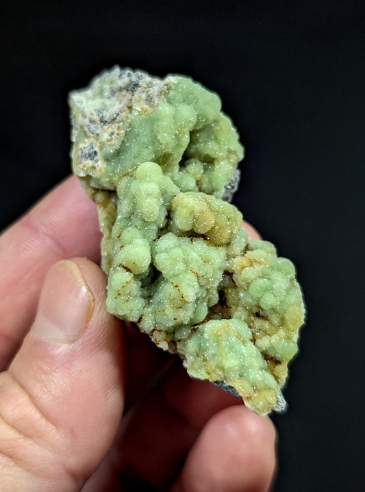 A hand holds a textured, light green mineral specimen with crystalline formations and subtle yellowish patches.