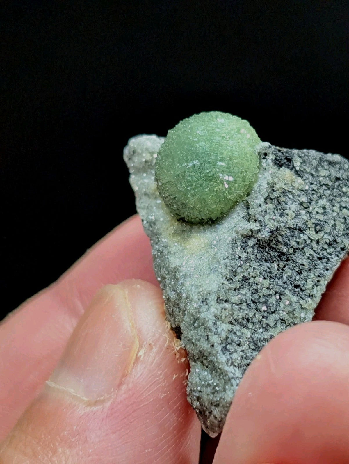 A green, spherical gemstone nestled atop a textured gray rock fragment.