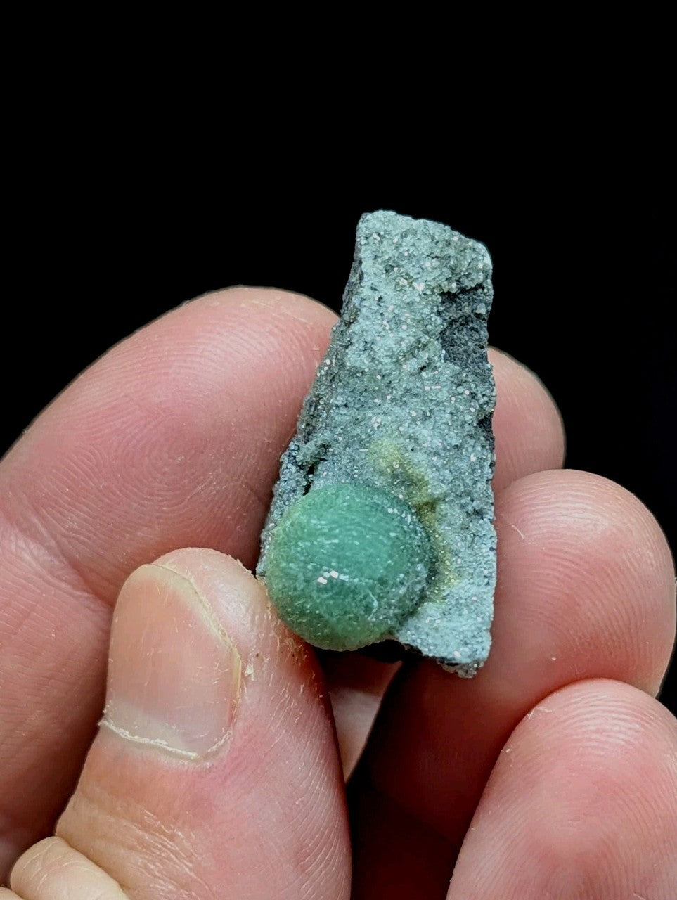 A vibrant green, translucent spherical mineral nestled within a rough-textured gray rock matrix.